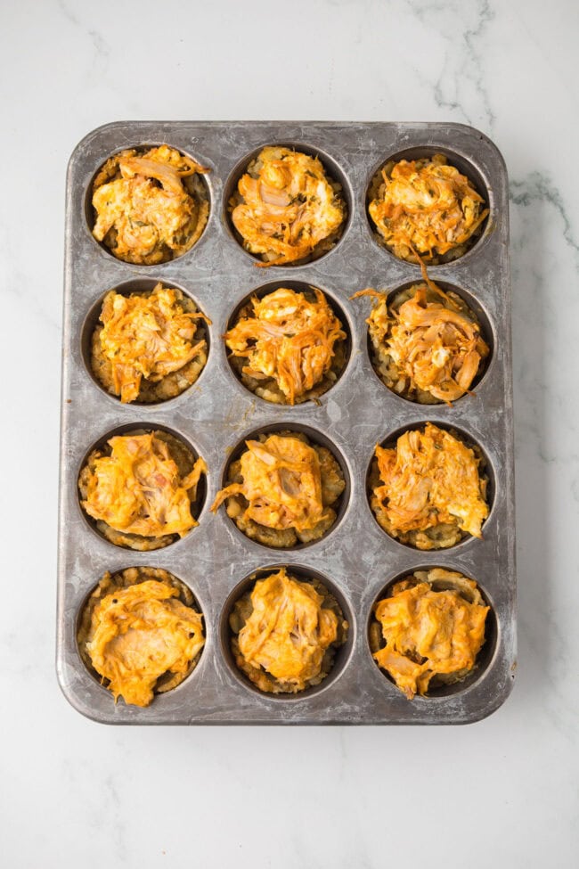 Twelve baked buffalo chicken tater tot cups in a muffin pan.