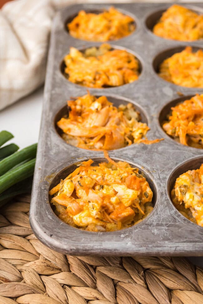 Close-up of baked buffalo chicken tater tot cups in a muffin pan on a woven placemat.