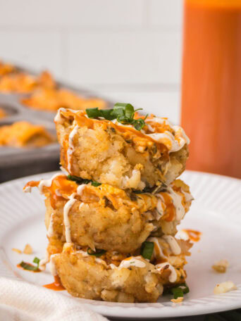 Three buffalo chicken tater tot cups stacked on a white plate with ranch drizzle and sliced green onions.
