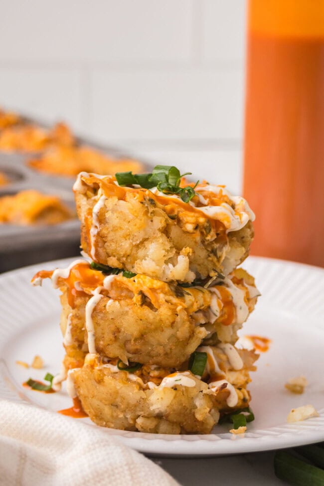 Three buffalo chicken tater tot cups stacked on a white plate with ranch drizzle and sliced green onions.