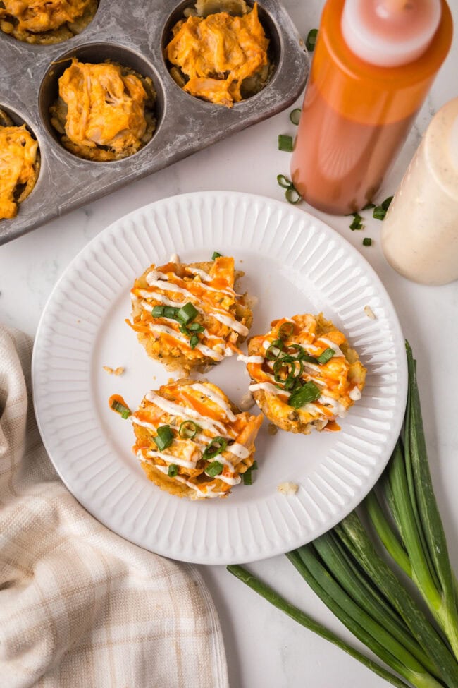 Overhead view of three buffalo chicken tater tot cups topped with ranch drizzle and sliced green onions on a white plate.
