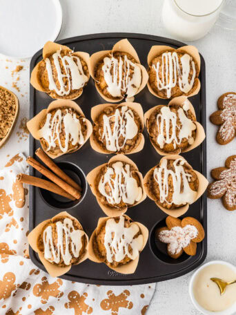 Overhead view of iced gingerbread muffins in a muffin pan with cinnamon sticks and gingerbread cookies.