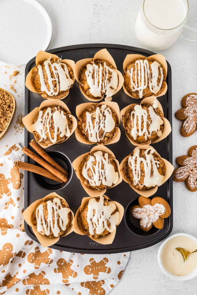 Overhead view of iced gingerbread muffins in a muffin pan with cinnamon sticks and gingerbread cookies.